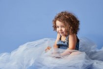 Adorable chica de pelo rizado sonriendo lindo sentado en tela delgada sobre fondo azul y mirando hacia otro lado - foto de stock
