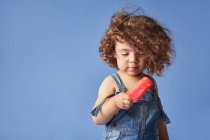 Unemotional thoughtful little girl standing with melting popsicle against blue background — Stock Photo