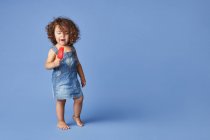 Pieno corpo di deliziata bambina in piedi con ghiacciolo sciogliente sullo sfondo blu — Foto stock