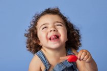 Смішна дівчина в джинсовому вбранні з кучерявим волоссям, що показує язик під час їжі солодкого морозива на синьому фоні — стокове фото