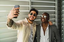 Счастливый бородатый этнический мужчина с татуировкой обнимающий черного партнера в стильном костюме и солнцезащитных очках, делая автопортрет на мобильном телефоне — стоковое фото