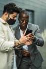 Elegante hombre afroamericano ejecutivo un traje cerca de socio étnico en la máscara de ver el teléfono celular en la calle de la ciudad - foto de stock