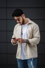 Фокусований бородатий етнічний чоловік в аксесуарах і модний одяг, що серфінгує Інтернет на мобільному телефоні на вулиці вдень — стокове фото