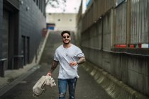 Jeune homme ethnique tatoué heureux dans des lunettes de soleil et jeans déchiré se promenant sur la passerelle entre les bâtiments urbains — Photo de stock