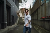Jeune homme ethnique tatoué heureux dans des lunettes de soleil et jeans déchiré se promenant sur la passerelle entre les bâtiments urbains — Photo de stock