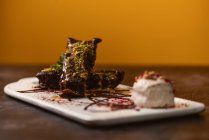 Tasty brownies covered with peanut butter and crunchy crushed pistachios near gelato scoop on plate with chocolate sauce on yellow background — Stock Photo