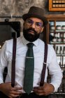 Crop bearded dandy ethnic male hairdresser in eyeglasses with mustache standing looking at camera in barbershop - foto de stock