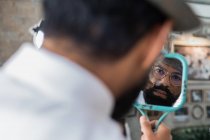 Back view of crop bearded dandy ethnic male hairdresser in eyeglasses reflecting in mirror while curling mustache in barbershop — Stock Photo