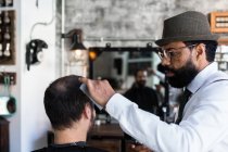 Стильный денди серьезный этнический мужчина парикмахер обрезать волосы взрослого клиента с электрическим клиппером в парикмахерской — стоковое фото