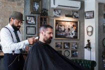 Vue latérale de barbier ethnique sérieux élégant dandy coupe les cheveux du client adulte avec tondeuse électrique dans le salon de coiffure — Photo de stock