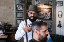 Стильний лайно серйозний етнічний чоловічий перукар обрізає волосся дорослого клієнта з електричним затискачем в перукарні — стокове фото
