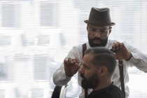 Sérieux mature barbu ethnique mâle barbier coupe les cheveux du client avec des ciseaux pendant la procédure de toilettage dans le salon de beauté — Photo de stock