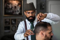 Serious mature bearded ethnic male barber trimming hair of client with scissors during grooming procedure in beauty salon - foto de stock