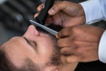 Crop anonymous ethnic male beauty master cutting mustache of bearded client using trimmer and comb in barbershop - foto de stock