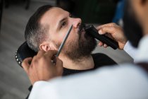 From above crop anonymous ethnic male beauty master cutting mustache of bearded client using trimmer and comb in barbershop - foto de stock