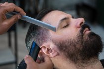 Crop anonymous ethnic male beauty master cutting mustache of bearded client using trimmer and comb in barbershop — Stock Photo