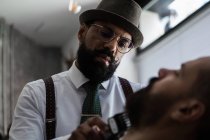 Stylish mature dandy ethnic male beauty master cutting mustache of bearded client using trimmer in barbershop - foto de stock