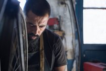 Концентрированный механик-мужчина с бородой и татуировками в перчатках, ремонтирующий велосипед в современной мастерской — стоковое фото