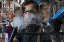 Uomo barbuto in grembiule con attrezzatura a spruzzo olio su ruota mentre ripara bici in garage — Foto stock