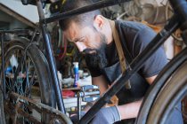 Concentrated male mechanic with beard and tattoos in gloves repairing bicycle in modern workshop — Stock Photo