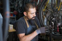 Homme adulte sérieux dans tablier et gants réparer roue de vélo dans le garage moderne — Photo de stock