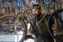 Homem adulto sério em avental e luvas de reparação roda de bicicleta na garagem moderna — Fotografia de Stock