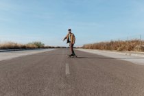 Молодий бородатий чоловічий фігурист у стильному одязі їде скейтборд уздовж асфальтової дороги в сільській місцевості — стокове фото
