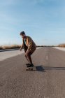 Full body young bearded male skater in stylish wear riding skateboard along asphalt road in countryside — Foto stock