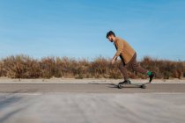 Side view full body young bearded male skater in stylish wear riding skateboard along asphalt road in countryside — Stock Photo