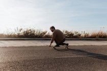 Ganzkörper junge bärtige männliche Skater in lässiger Kleidung Durchführung Trick berühren Boden während der Fahrt auf Asphaltstraße — Stockfoto