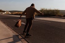 Full body back view of unrecognizable male skater in trendy clothes with leather bag riding skateboard along asphalt road — Foto stock