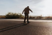 Full body back view of anonymous male skater in stylish wear riding skateboard along asphalt road in countryside - foto de stock
