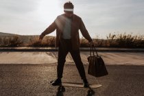 Full body serious young bearded male skater in trendy clothes with leather bag riding skateboard along asphalt road — Fotografia de Stock