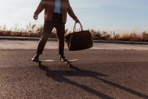 Cropped unrecognizable male skater in trendy clothes with leather bag riding skateboard along asphalt road — Fotografia de Stock