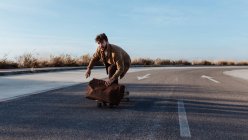 Full body serious young bearded male skater in trendy clothes with leather bag riding skateboard along asphalt road — Fotografia de Stock