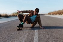 Full body young bearded male skater in casual clothes performing trick touching ground while riding on asphalt road — Foto stock