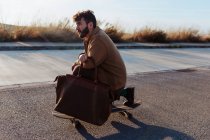 Side view of full body serious young bearded male skater in trendy clothes with leather bag riding skateboard along asphalt road — Foto stock