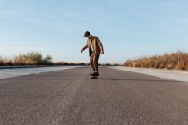 Full body young bearded male skater in stylish wear riding skateboard along asphalt road in countryside — Stock Photo