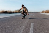 Full body young bearded male skater in casual clothes performing trick touching ground while riding on asphalt road — Fotografia de Stock