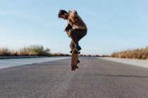 Полное тело молодой бородатый фигурист в повседневной одежде прыжки во время выполнения kickflip на скейтборде на асфальтовой дороге — стоковое фото