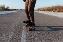 Обрезанный анонимный мужчина-фигурист в стильной одежде на скейтборде вдоль асфальтовой дороги в сельской местности — стоковое фото