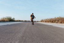 Молодий бородатий чоловічий фігурист у стильному одязі їде скейтборд уздовж асфальтової дороги в сільській місцевості — стокове фото
