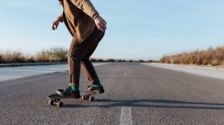 Cropped anonymous male skater in stylish wear riding skateboard along asphalt road in countryside — Stock Photo