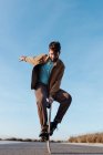 Full body young bearded male skater standing on edge of skateboard keeping balance while performing trick on asphalt road with hand raised and looking down — Stock Photo