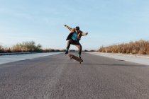 Full body young bearded skater in casual outfit jumping while performing kickflip on skateboard on asphalt road — Stock Photo