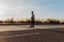 Full body young bearded male riding skateboard with bag and jacket in hand along pavement near asphalt road — Fotografia de Stock