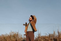 Улыбающийся молодой бородатый мужчина-фигурист в повседневной одежде стоит рядом с травой и забором со скейтбордом и трогательными волосами, отворачиваясь — стоковое фото