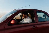 Bearded young male driver in white shirt sitting in car parked on roadside in countryside - foto de stock