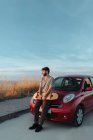 Full body thoughtful young bearded male skater in casual clothes leaning on hood of car parked on roadside with skateboard in hands and looking away - foto de stock