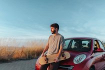Thoughtful young bearded male skater in casual clothes leaning on hood of car parked on roadside with skateboard in hands and looking away — Fotografia de Stock
